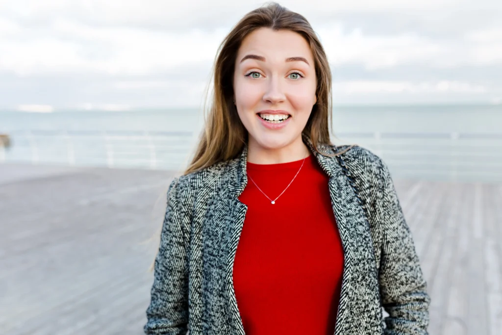 happy smiling long haired woman with big blue eyes red shirt grey coat walks near sea
